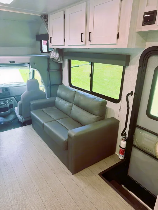 Clean RV rental interior with a gray sofa, white cabinets, light wood floor, and front cab view. Fire extinguisher near entry.. East to West Entrada 2023