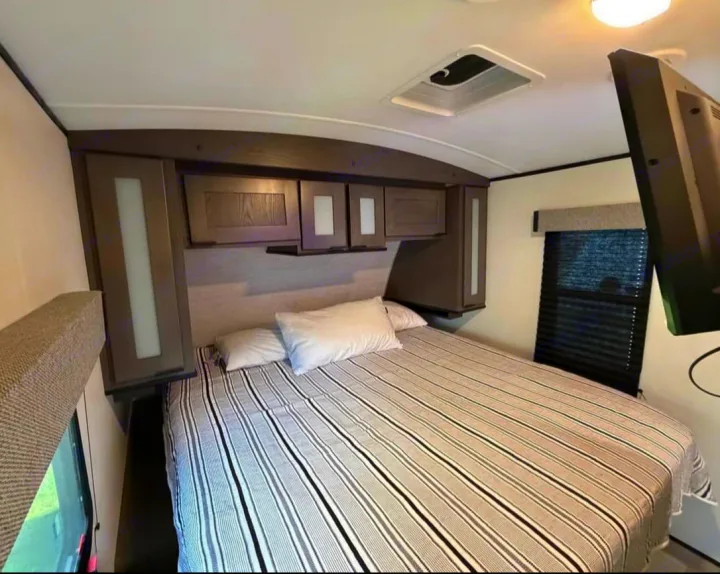 Cozy rv rental bedroom with a striped bed, overhead cabinets, and a wall-mounted TV.. Cruiser RV MPG Ultra Lite 2021