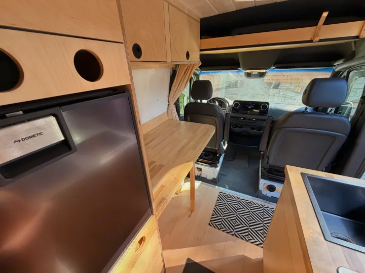 Inside an RV rental conversion van: light wood cabinets, Dometic fridge, sink, and front seats for a cozy camper experience.. Mercedes-Benz Sprinter 2019
