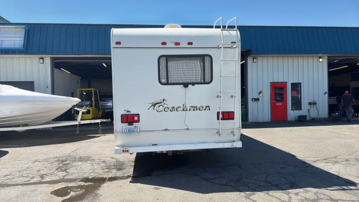 Rear of a white Coachmen RV rental with ladder, license plate CFR6022, parked outside a busy service garage with a boat nearby.. Coachmen Leprechaun 2003