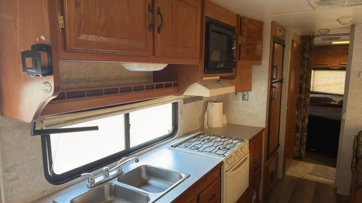 RV rental interior with a compact kitchen: dual sink, stove, microwave, cabinets, and a glimpse into the sleeping area.. Coachmen Leprechaun 2003