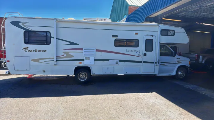 This white Coachmen Leprechaun Class C RV rental is parked near a service bay, showing its full side profile.. Coachmen Leprechaun 2003