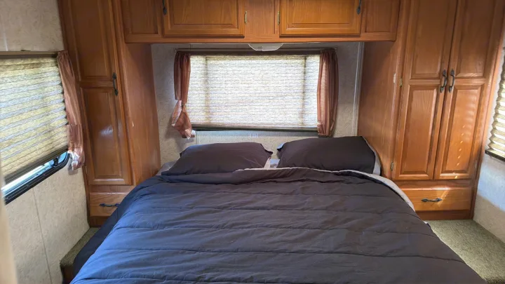 An RV rental bedroom with a neatly made bed, dark bedding, a window with closed blinds, and built-in wooden cabinets.. Coachmen Leprechaun 2003