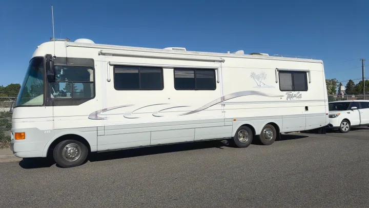Cream-colored Tropical RV rental, a large motorhome with a small white car in tow, parked under a clear blue sky.. Intermountain Rv TROPICAL 1997