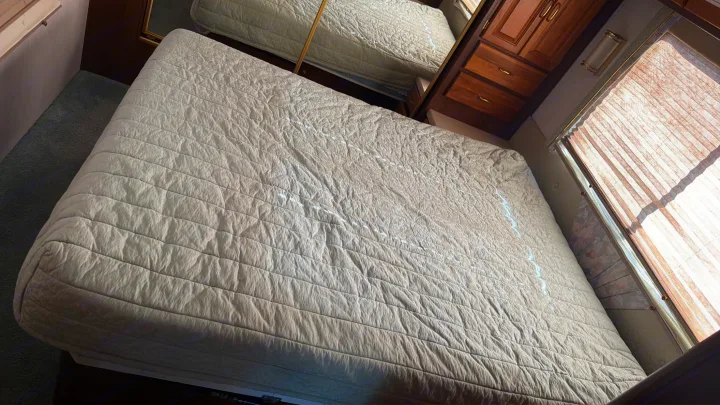Inside an RV rental, showing a bed with quilted bedding, wood cabinets, and a sunlit window with blinds.. Intermountain Rv TROPICAL 1997