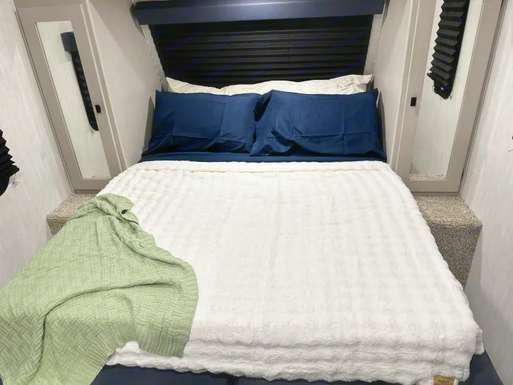 Cozy RV rental bedroom with a neatly made bed, featuring navy sheets, a white fluffy blanket, and a green throw.. Coachmen RMT18R 2024