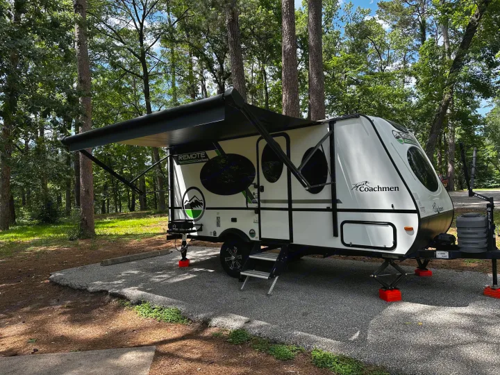 White Coachmen rv rental travel trailer set up on a gravel campsite amidst tall forest trees, awning extended.. Coachmen RMT18R 2024