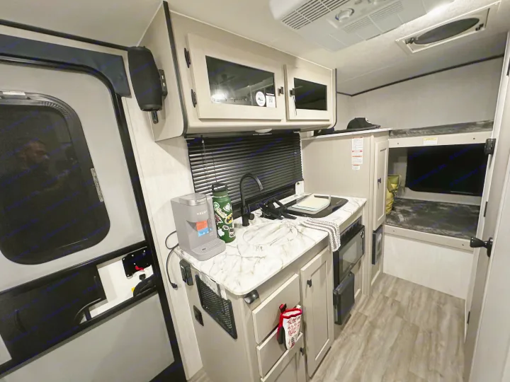 This RV rental interior showcases a compact kitchen with a Keurig, sink, & stove, alongside a cozy bunk bed area.. Coachmen RMT18R 2024