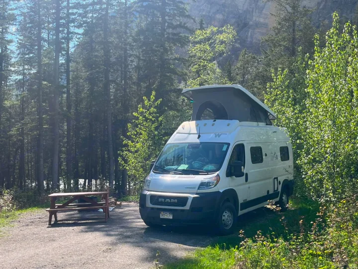 White RAM camper van (rv rental) with pop-top roof at a forest campsite with picnic table, surrounded by trees and mountains.. Winnebago Solis 2023