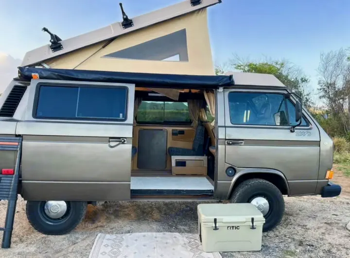 Vintage VW camper van with pop-top roof up & side door open, perfect for an RV rental road trip. Cooler & mat outside.. Volkswagen Westfalia 1985