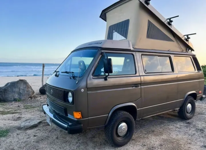 Brown Westfalia pop-top camper van on a sandy beach with ocean waves. An ideal RV rental for outdoor adventures.. Volkswagen Westfalia 1985