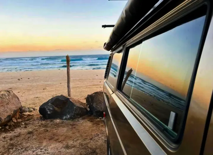 Enjoy coastal views from your RV rental. A camper van parked on a sandy beach with ocean waves visible, reflecting in the window.. Volkswagen Westfalia 1985