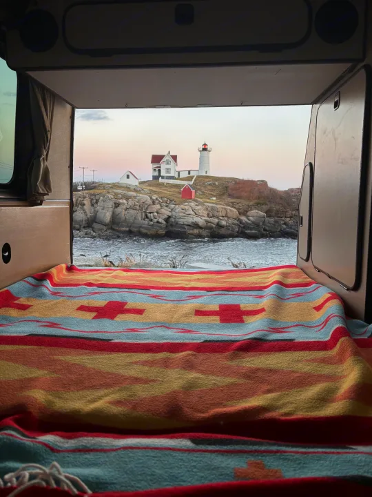 Cozy rv rental campervan bed with a vibrant blanket, offering a stunning ocean view of Nubble Lighthouse. Ideal for scenic travel.. Volkswagen Westfalia 1985