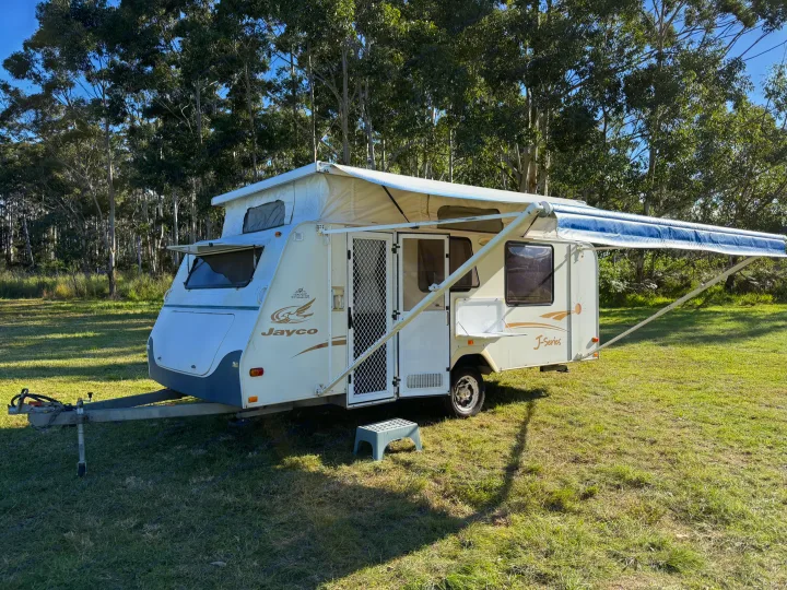 White Jayco J-Series pop-top rv rental camper trailer with awning extended, parked on a grassy field among tall trees.. Jayco J Series 2008
