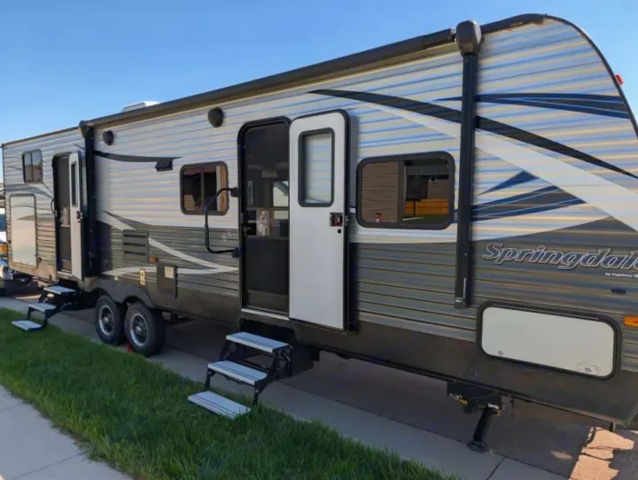 Side view of a Springdale RV rental travel trailer with open doors, extended steps, parked on pavement by grass under blue sky.. Keystone RV Springdale 2019