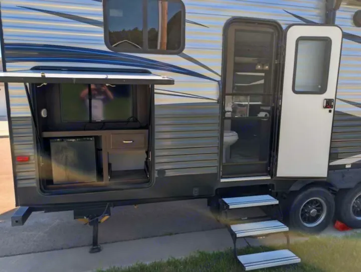 Side view of an RV rental travel trailer featuring an outdoor entertainment center with TV, mini-fridge, and open main door with steps.. Keystone RV Springdale 2019