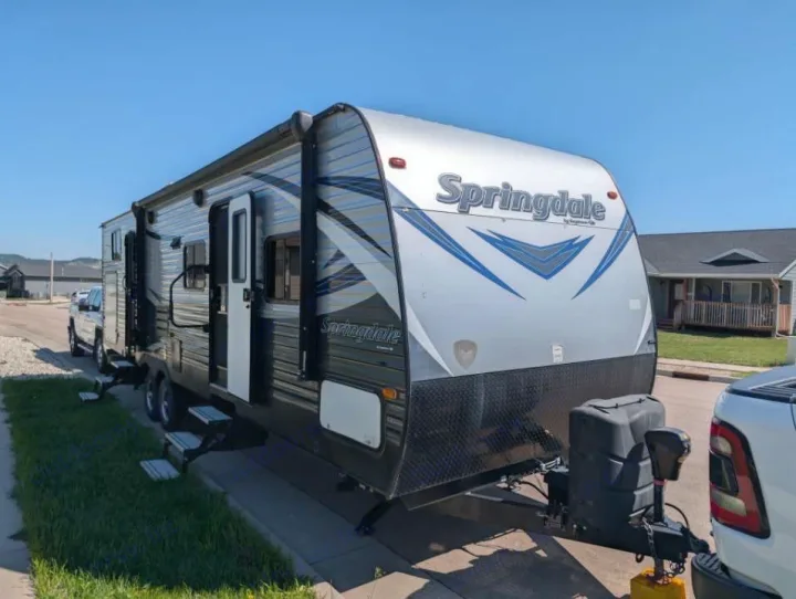 This Springdale travel trailer (RV rental) is shown hitched to a white pickup truck on a sunny residential street.. Keystone RV Springdale 2019