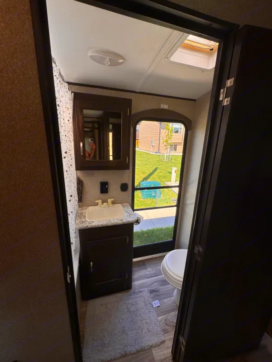 Compact RV rental bathroom with a sink, mirror, toilet, and window offering an outdoor view. Features a patterned shower curtain.. Keystone RV Springdale 2019