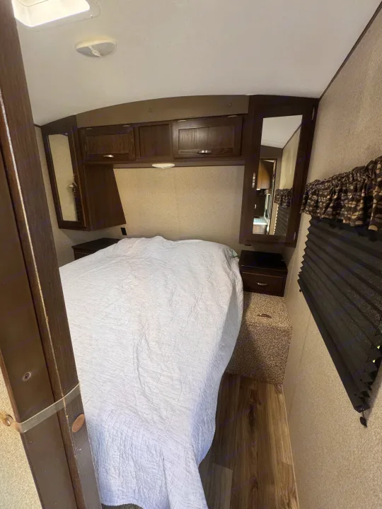 Inside an RV rental, this cozy bedroom features a white quilted bed, overhead cabinets, bedside tables, and pleated window blinds.. Keystone RV Springdale 2019