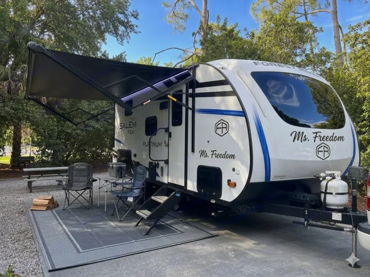 White travel trailer RV rental at a sunny campsite, featuring an extended awning, outdoor seating, and a mat.. Forest River Salem FSX 2025