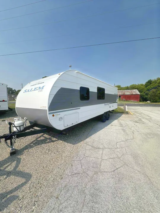 Forest River Salem FSX travel trailer for rv rental. White and gray, parked on gravel, showing propane tank, windows, and a red barn.. Forest River Salem FSX 266BHLE 2026