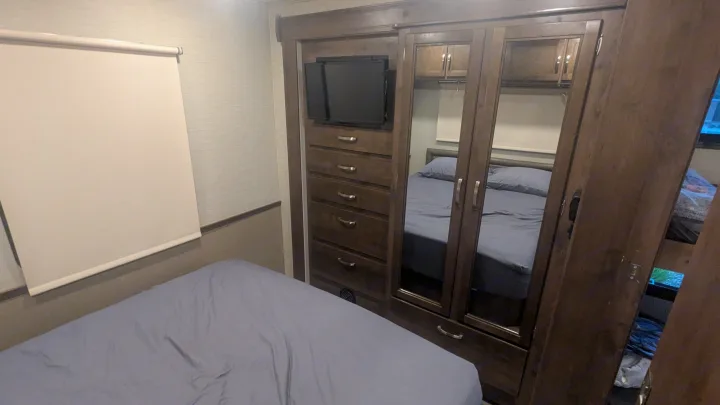 Inside an RV rental, a compact bedroom with a bed, window blind, and built-in wood unit with TV & mirrored closet.. Jayco Alante 2020