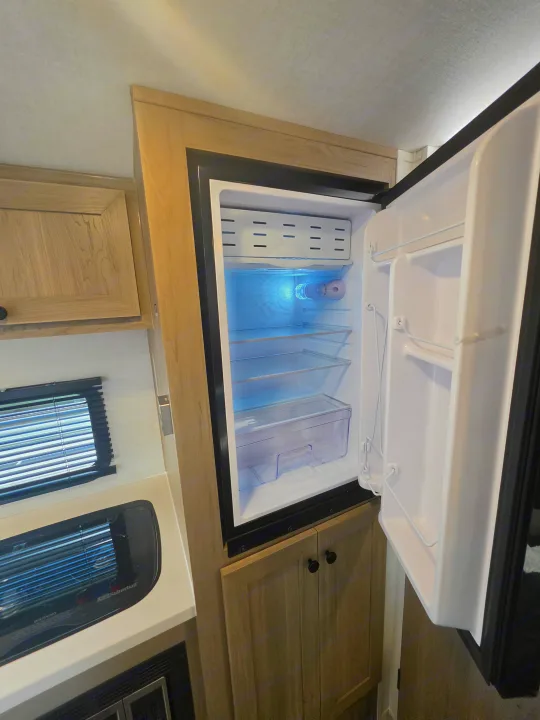 RV rental interior: an open, empty refrigerator with blue light, beside light wood cabinets, a window, and appliances.. Forest River Rockwood Geo Pro 2026
