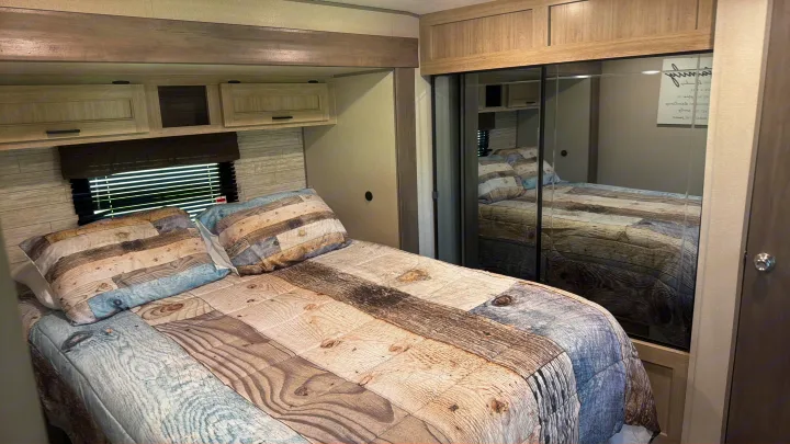 This RV rental bedroom features a comfy bed with unique wood-patterned bedding, overhead storage, and a mirrored closet.. Coachman Catalina, Legacy edition 343BHTS 2024