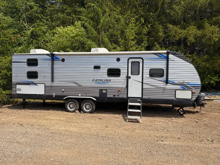 Large travel trailer RV rental, grey with blue accents, parked on gravel against a lush evergreen forest.. Coachmen Other 2021