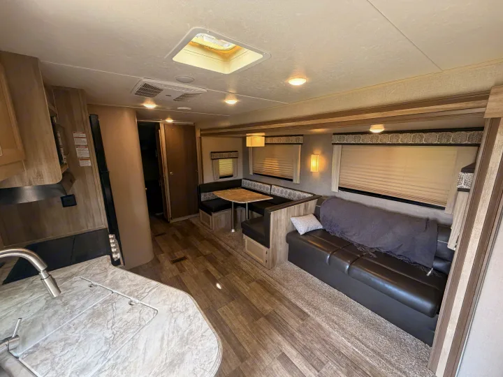 A bright RV rental interior showing a kitchen, dinette with seating for four, and a cozy sofa with a blanket, on wood-look flooring.. Coachmen Other 2021