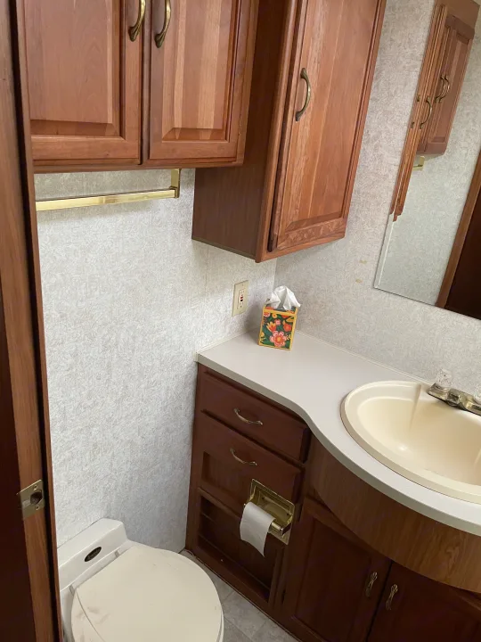 Inside an RV rental: a compact bathroom with wood cabinets, a sink, toilet, and mirror, offering essential amenities.. Winnebago Chieftain 1999