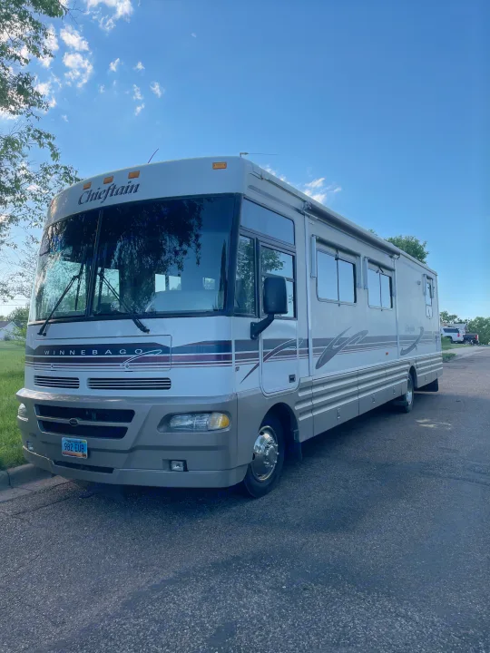 Front and side view of a large, light-colored Winnebago Chieftain RV rental, parked on asphalt under a bright blue sky.. Winnebago Chieftain 1999