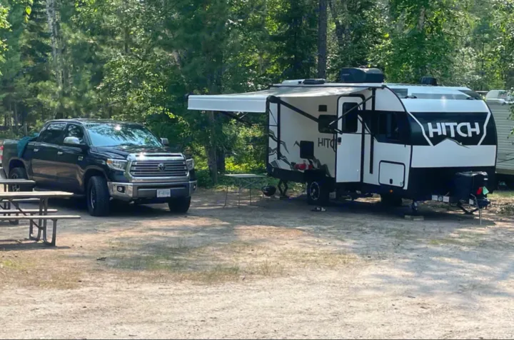 A black pickup truck and a white/black HITCH travel trailer (RV rental) are parked at a wooded campsite.. Cruiser RV Hitch 2022