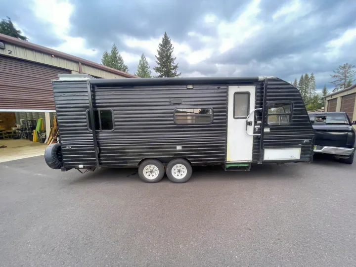 A black travel trailer, perfect for an rv rental, is parked near storage units on a cloudy day.. Fleetwood Prowler 1996