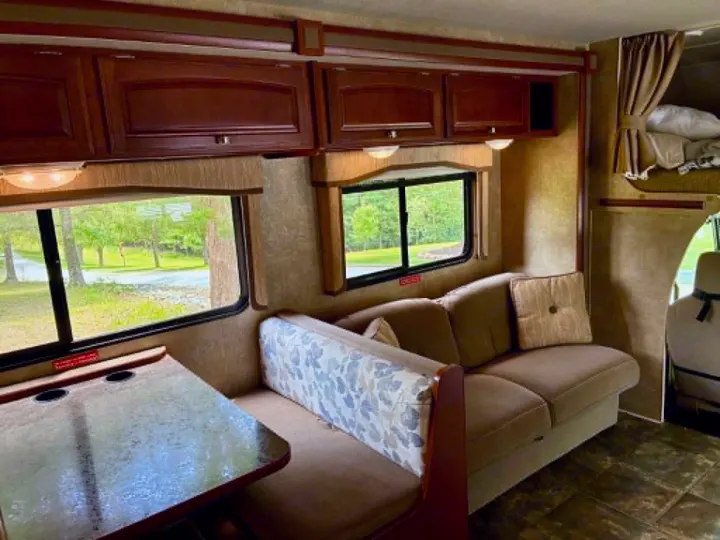 RV rental interior: cozy dinette, sofa, and bunk bed area with wood cabinets. Large windows show a scenic outdoor view.. Winnebago Chalet 2014