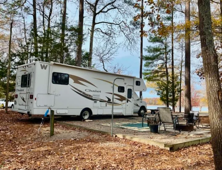 White RV rental parked at a serene lakeside campground, surrounded by autumn trees and a patio seating area with a fire pit.. Winnebago Chalet 2014