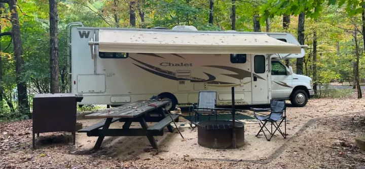 An RV rental is parked at a serene wooded campsite with a picnic table, fire pit, and chairs. Ready for camping!. Winnebago Chalet 2014