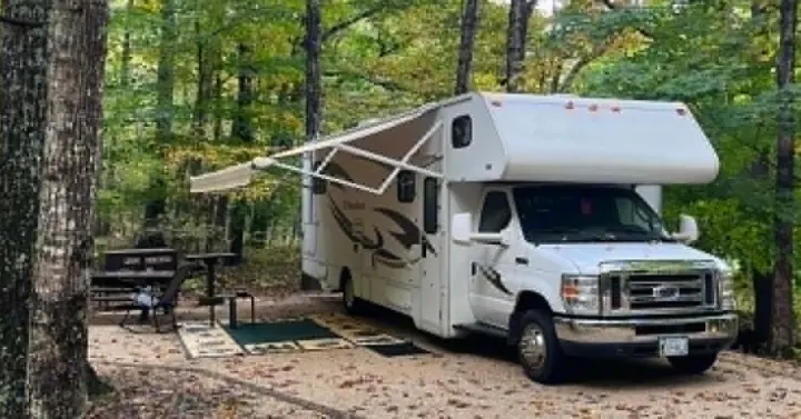 White RV rental with awning extended at a rustic campsite, featuring a picnic table and chairs for outdoor relaxation.. Winnebago Chalet 2014