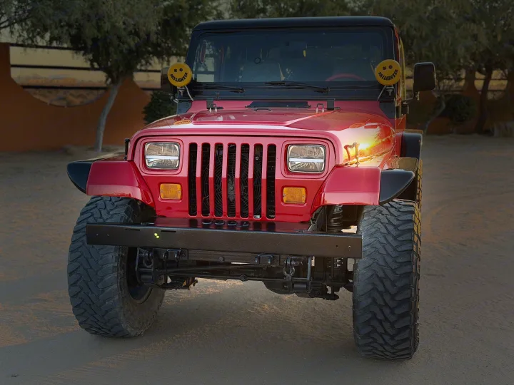 Front view: Rugged red Jeep with large tires & KC Daylighter lights, parked outdoors. An adventurous vehicle beyond RV rental options.. Entegra Coach Qwest 2020