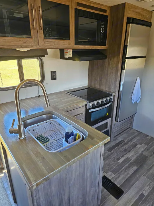 Inside an RV rental, this kitchen features wood-look counters, a stainless steel sink, stove, microwave, and refrigerator.. KZ Sportsman 2022