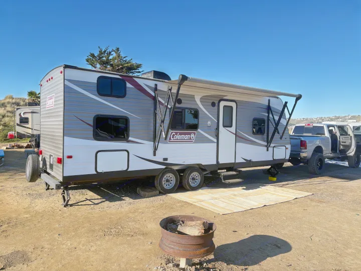 A Coleman travel trailer RV rental at a sunny campsite, hooked to a pickup truck, with an extended awning and fire pit.. Coleman Lantern 2018