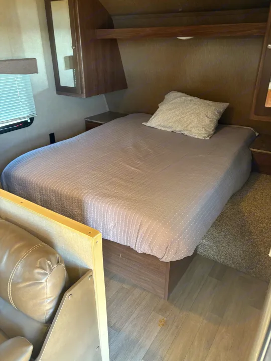 RV rental bedroom view featuring a neatly made bed with a grey quilted comforter, wooden overhead cabinets, and a window with blinds.. Coleman Lantern 2018