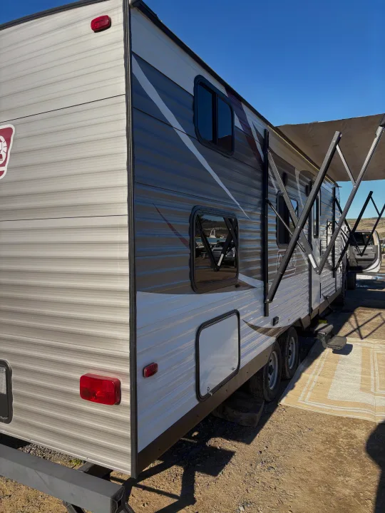 An RV rental features an extended awning & modern gray/white design, parked on a sunny camping site.. Coleman Lantern 2018