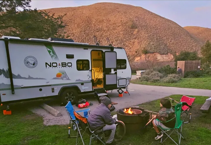 Two people by a campfire with an RV rental at dusk, parked at a scenic campsite with mountains.. No Boundaries 19 2022