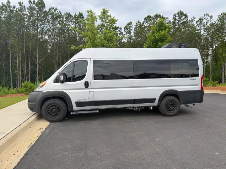 White Airstream Rangeline rv rental camper van, parked on asphalt. Features side steps & roof AC, surrounded by trees.. Airstream Airstream Class B 2023