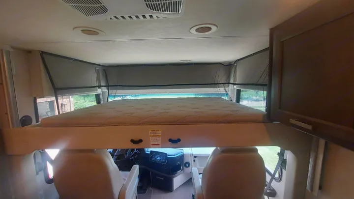 This RV rental interior features an overhead bunk bed with safety netting, situated above the driver's and passenger's seats.. Thor Palazzo 2023