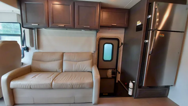 RV rental interior featuring a beige sofa, overhead cabinets, an entry door, a stainless steel fridge, and a fire extinguisher.. Thor Palazzo 2023