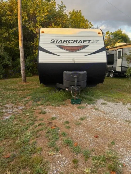 A white and black StarCraft travel trailer RV rental is parked on a grassy, gravel lot with trees in the background.. Starcraft Autumn Ridge Outfitter 2018
