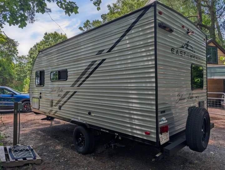 Grey East West Silver Lake RV rental, a travel trailer, parked outdoors on gravel. Features include windows, spare tire, & NC license plate.. East to West Silver Lake 2023