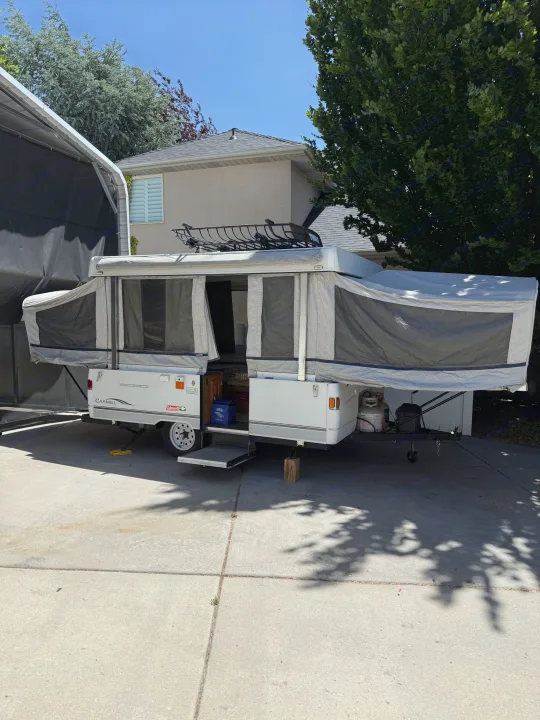 White and gray pop-up camper RV rental, fully extended with a roof rack, parked on a sunny paved driveway.. Coleman Grand Tour 2003